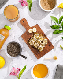 Flat-lay healthy breakfast on marble countertop: banana slices on chia-seed toast on a wooden board, bowl of chia seeds, nut butter, orange juice, honey dipper and flowers.