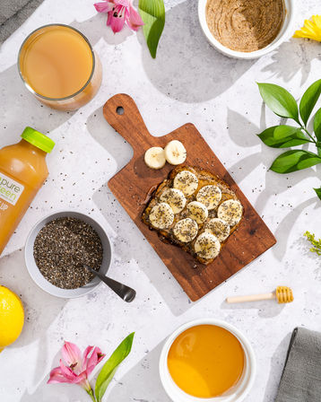 Flat-lay healthy breakfast on marble countertop: banana slices on chia-seed toast on a wooden board, bowl of chia seeds, nut butter, orange juice, honey dipper and flowers.