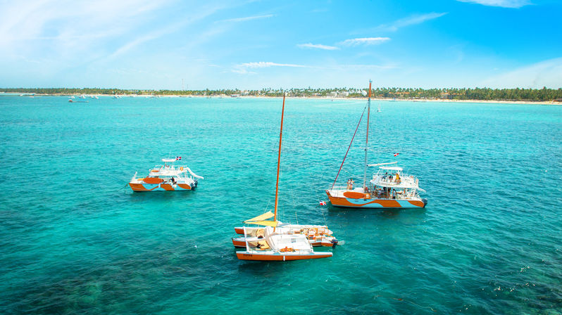 Three orange-and-white catamarans anchored in clear turquoise tropical water near a palm-lined sandy coastline under a bright blue sky