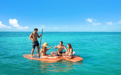 Group of four on orange paddleboards in clear turquoise tropical sea under a sunny blue sky, one standing with a paddle while others sit and share drinks.