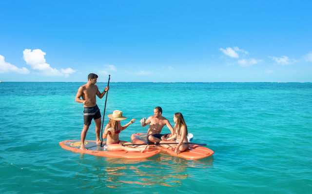 Group of four on orange paddleboards in clear turquoise tropical sea under a sunny blue sky, one standing with a paddle while others sit and share drinks.