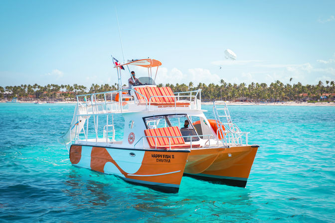 Orange-and-white catamaran with striped cushions floating in crystal turquoise tropical water near a palm-lined beach under a sunny sky with a parasailer.
