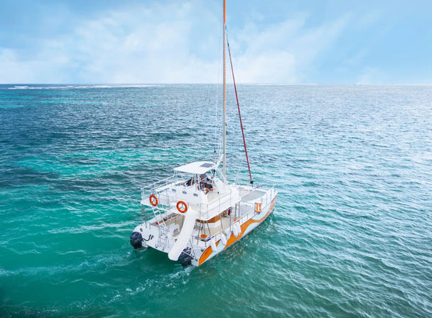 Aerial view of a white-and-orange catamaran sailboat with a stern slide and life rings floating on turquoise tropical ocean under a bright blue sky.