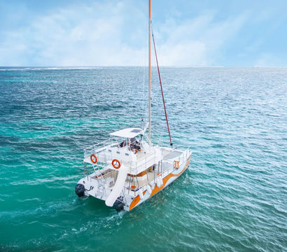 Aerial view of a white-and-orange catamaran sailboat with a stern slide and life rings floating on turquoise tropical ocean under a bright blue sky.