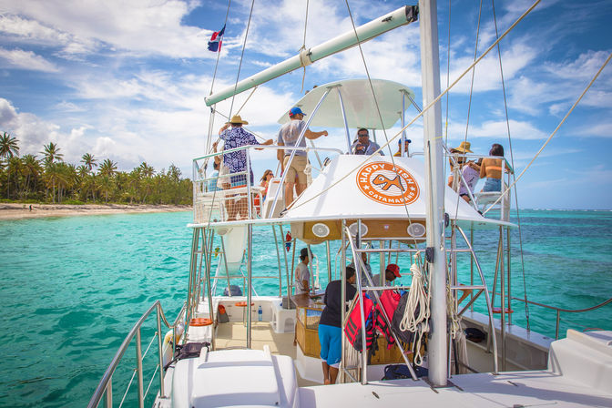 Passengers enjoying a sunny catamaran cruise in turquoise tropical waters near a palm-lined beach and blue sky.