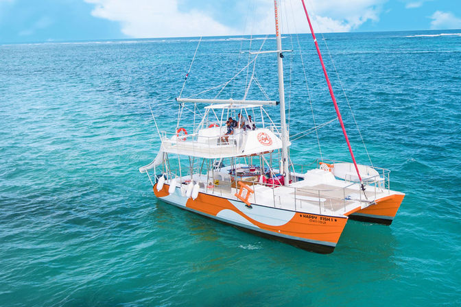 Orange-and-white catamaran cruising on clear turquoise tropical ocean under a blue sky, passengers relaxing on deck during a sightseeing boat tour.