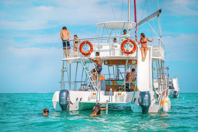 Tropical catamaran excursion in turquoise Caribbean waters with people climbing ladders, sliding from the upper deck into the sea, and swimming under a bright blue sky.
