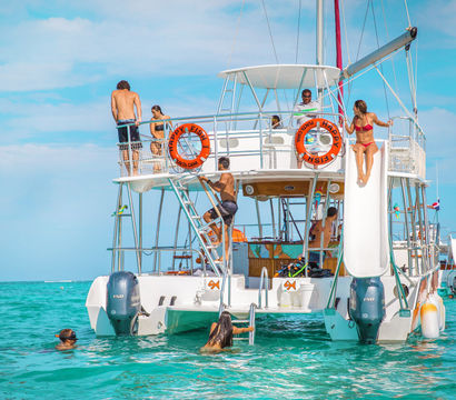 Tropical catamaran excursion in turquoise Caribbean waters with people climbing ladders, sliding from the upper deck into the sea, and swimming under a bright blue sky.