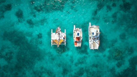 Aerial shot of three anchored catamarans and sailboats lined up over crystal-clear turquoise tropical water with visible coral patches.