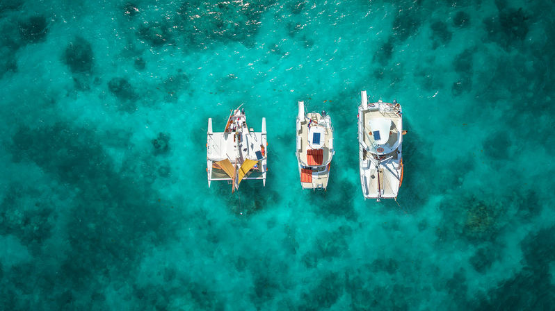 Aerial shot of three anchored catamarans and sailboats lined up over crystal-clear turquoise tropical water with visible coral patches.
