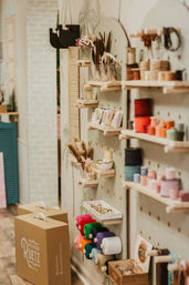 Cozy craft shop interior with pegboard display of colorful yarn spools, ribbons, jars of feathers, beads and small shelves of DIY supplies beside brown carry boxes on a wooden floor