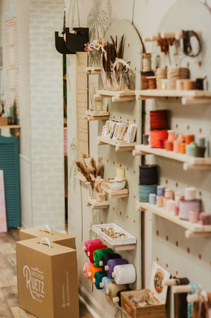 Cozy craft shop interior with pegboard display of colorful yarn spools, ribbons, jars of feathers, beads and small shelves of DIY supplies beside brown carry boxes on a wooden floor