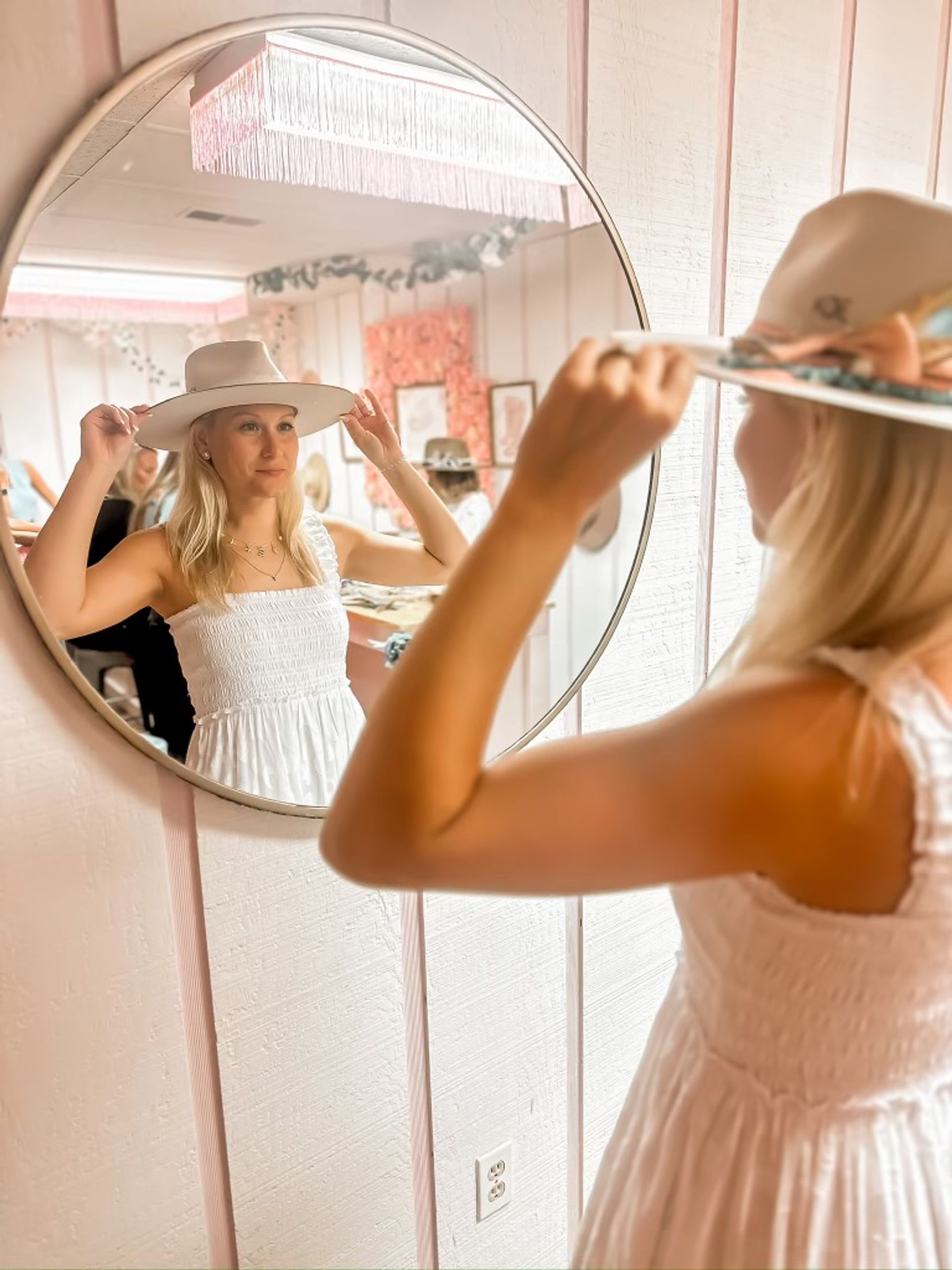 Person in white sundress trying on a wide-brim hat in a pink boutique dressing area, reflected in a round mirror