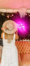 Person seen from behind in a white sundress and beige boho hat posing at an indoor photo wall with faux greenery and roses, glowing pink neon sign, crystal chandelier and tufted peach sofa.