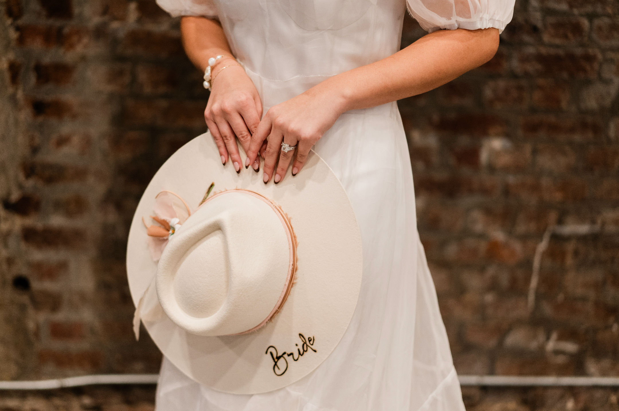 Bride in a white dress holding a wide-brimmed hat labeled 'Bride', showcasing an engagement ring against a rustic brick wall backdrop
