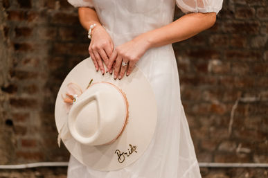 Bride in a white dress holding a wide-brimmed hat labeled 'Bride', showcasing an engagement ring against a rustic brick wall backdrop