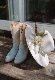 Two-tone light blue suede cowboy boots beside a white western hat decorated with silk flowers, greenery and a feather, resting on a weathered wooden bench outdoors