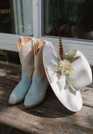 Two-tone light blue suede cowboy boots beside a white western hat decorated with silk flowers, greenery and a feather, resting on a weathered wooden bench outdoors