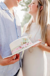 Sunlit outdoor shot of a smiling couple holding a white brimmed hat wrapped in pink floral ribbon with a small 'Charleston' postcard, soft romantic vibe