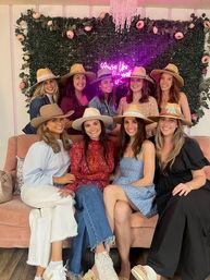 Nine smiling women wearing straw and felt hats, seated on a pink velvet sofa in front of a flower-adorned greenery wall with a neon sign — playful indoor boutique-style group photo.