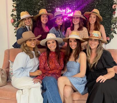 Nine smiling women wearing straw and felt hats, seated on a pink velvet sofa in front of a flower-adorned greenery wall with a neon sign — playful indoor boutique-style group photo.