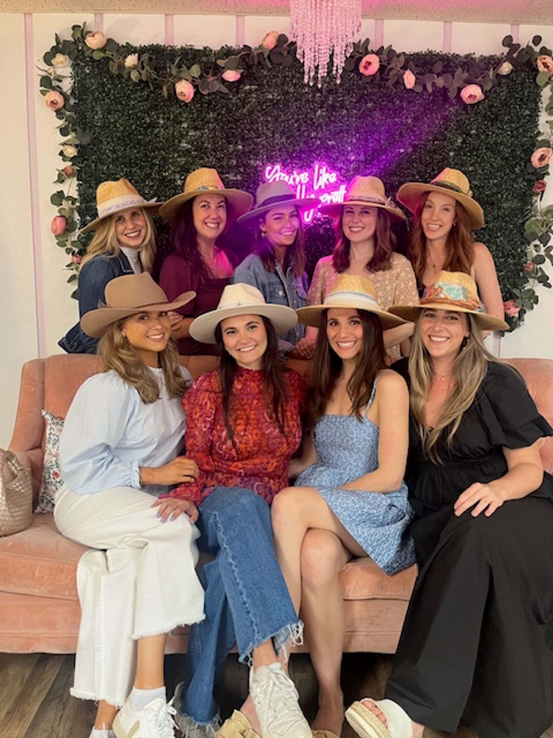 Nine smiling women wearing straw and felt hats, seated on a pink velvet sofa in front of a flower-adorned greenery wall with a neon sign — playful indoor boutique-style group photo.