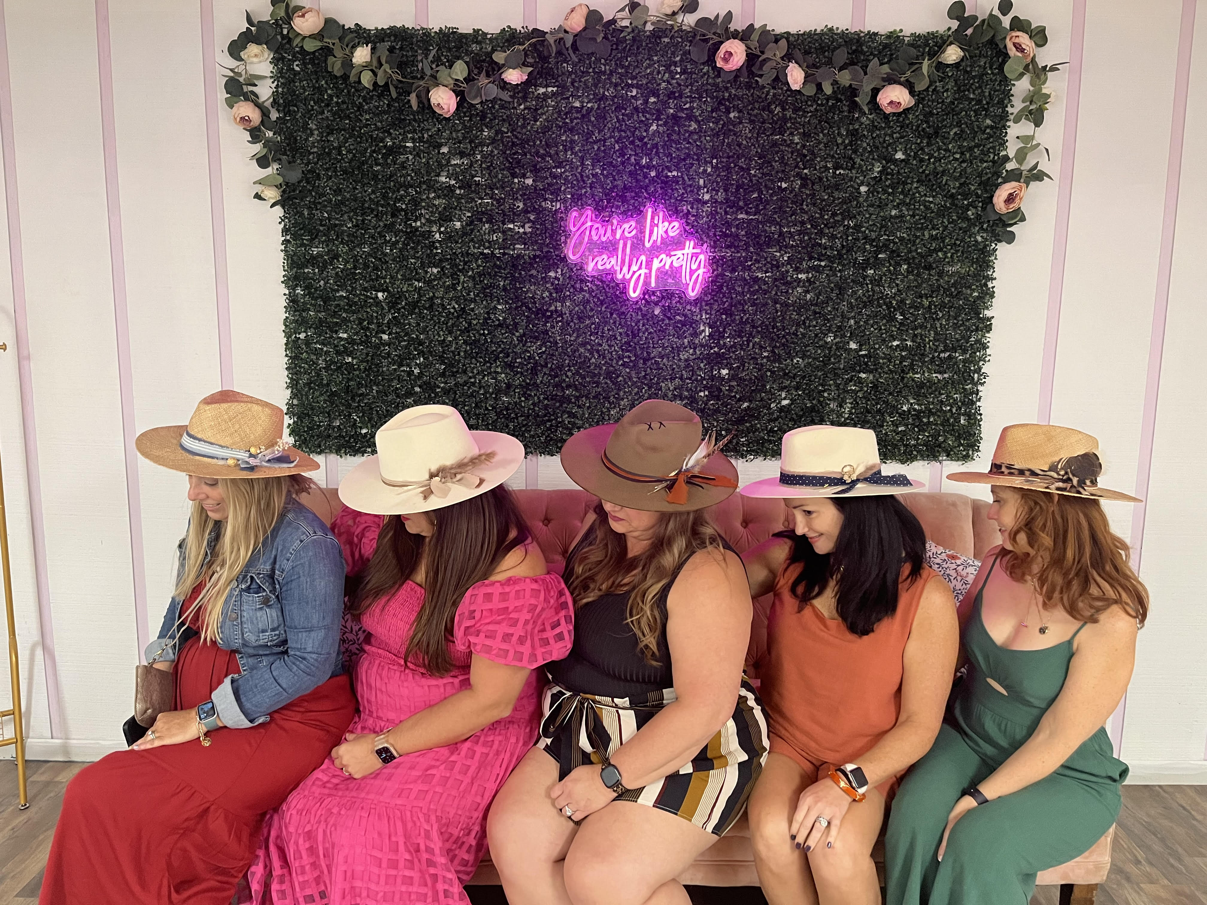Five women wearing wide-brimmed summer hats and colorful dresses sit on a blush sofa against a green foliage wall with roses and a pink neon sign reading "You're like really pretty".