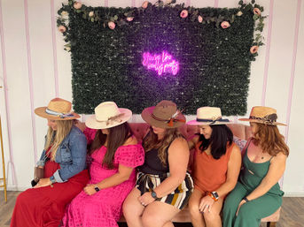 Five women wearing wide-brimmed summer hats and colorful dresses sit on a blush sofa against a green foliage wall with roses and a pink neon sign reading "You're like really pretty".