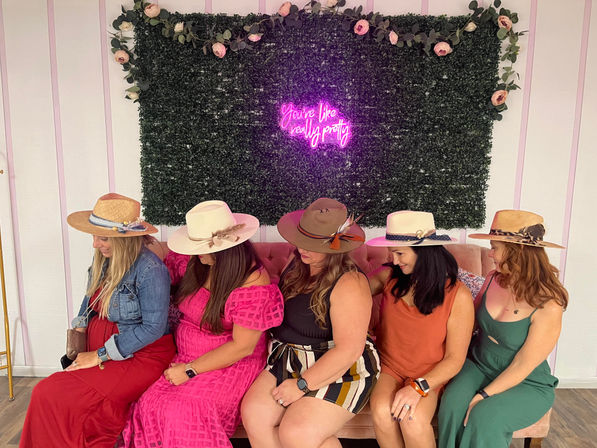 Five women wearing wide-brimmed summer hats and colorful dresses sit on a blush sofa against a green foliage wall with roses and a pink neon sign reading "You're like really pretty".