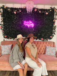 Two smiling women in wide-brim hats seated on a blush velvet sofa against a greenery wall with roses, a pink neon sign reading 'You're like really pretty' and a crystal chandelier — boutique photo lounge backdrop