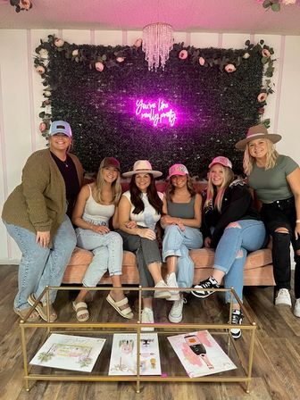 Six women posing on a pink velvet couch in a chic indoor party lounge with a greenery-and-rose backdrop, pink neon sign, crystal chandelier, casual hats and jeans, and a glass coffee table with prints — playful celebration photo.