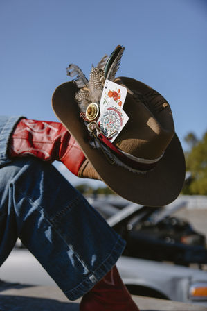 Brown felt cowboy hat with feathers, vintage pin and floral playing cards tucked in the band, styled with red leather cowboy boots and denim jeans in a sunny outdoor western-style scene