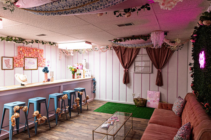 Cozy pink floral-themed studio interior with a long wooden bar, blue stools adorned with dried flowers, velvet tufted sofa, glass coffee table, neon wall sign and hanging floral garlands.