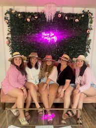 Five friends in summer hats seated on a pink velvet sofa at a floral wall photo booth with pink neon sign 'You're like really pretty' and a crystal chandelier.