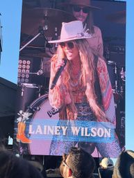 Outdoor concert giant screen showing a country singer in a wide‑brim hat and sunglasses holding a mic, drum kit behind, colorful boho outfit and crowd in foreground.