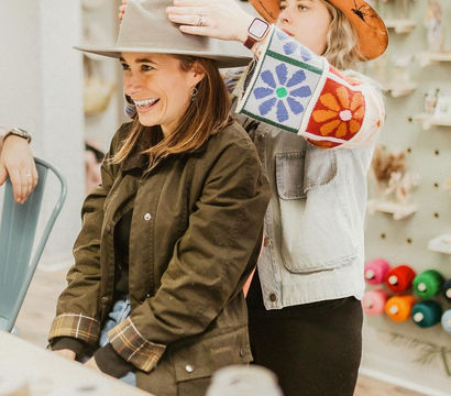 Smiling woman tries on a wide-brim felt hat in a cozy boutique while a staffer in a colorful floral-sleeve jacket adjusts it, with yarn spools and craft supplies on the wall behind them.