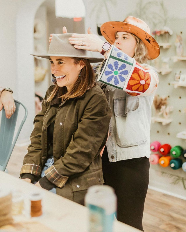 Smiling woman tries on a wide-brim felt hat in a cozy boutique while a staffer in a colorful floral-sleeve jacket adjusts it, with yarn spools and craft supplies on the wall behind them.