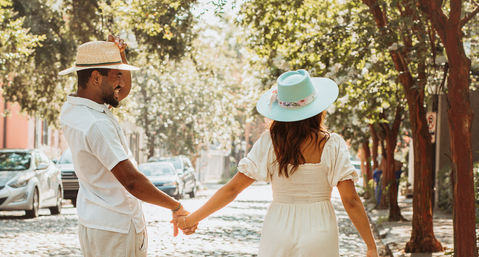 Couple holding hands strolling down a sunlit, tree-lined cobblestone street — woman in a light summer dress with a teal wide-brimmed hat and man in a straw hat and white shirt smiling and tipping his hat, parked cars along the block.