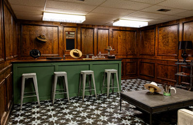 Vintage wood-paneled basement bar with a green counter and four metal stools, star-patterned black-and-white tile floor, cowboy hats displayed on walls and table, low fluorescent ceiling lights