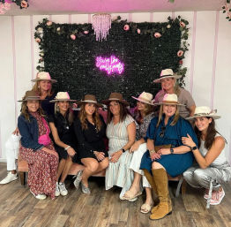 Nine friends wearing wide-brim hats pose on a pink velvet sofa in a boutique-style indoor photo area with floral greenery, crystal chandelier, and pink neon sign.