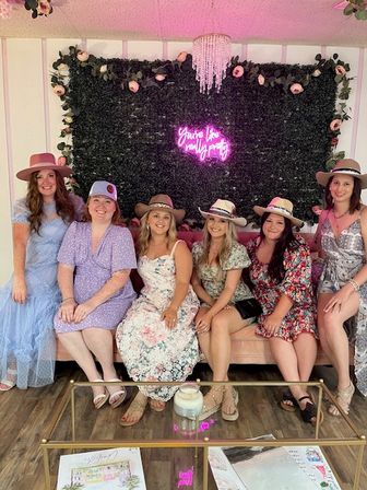 Six women in floral dresses and hats seated on a pink velvet sofa in an indoor photo lounge with a rose‑garlanded greenery backdrop, pink neon sign, chandelier and glass coffee table — cheerful group celebration.