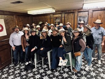 Group of about 18 adults in cowboy hats smiling and posing in a wood-paneled room with star-patterned tile floor — casual western-themed indoor group photo