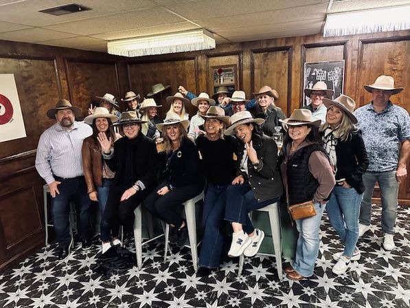 Group of about 18 adults in cowboy hats smiling and posing in a wood-paneled room with star-patterned tile floor — casual western-themed indoor group photo