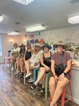 Six women wearing wide-brim hats sit on colorful stools in a bright hat boutique studio with hat displays and skyline wall art