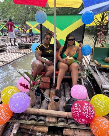 Two women lounging on a colorful bamboo raft in Jamaica, wearing sunglasses and enjoying a fresh coconut, surrounded by birthday balloons, umbrellas and Jamaican flag decor on a tropical river.