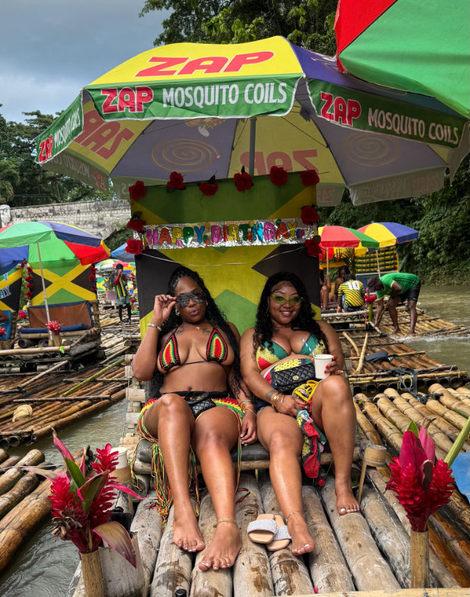 Two women in colorful reggae-style bikinis relaxing and smiling on a decorated bamboo raft under a large umbrella, surrounded by Jamaican-flag colors, tropical flowers and other rafts on a Jamaican river.