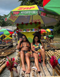 Two women in colorful reggae-style bikinis relaxing and smiling on a decorated bamboo raft under a large umbrella, surrounded by Jamaican-flag colors, tropical flowers and other rafts on a Jamaican river.