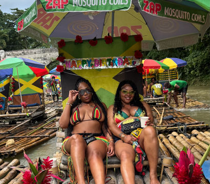 Two women in colorful reggae-style bikinis relaxing and smiling on a decorated bamboo raft under a large umbrella, surrounded by Jamaican-flag colors, tropical flowers and other rafts on a Jamaican river.