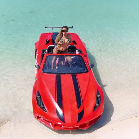 Woman in a patterned bikini posing in a red sports-car–style boat beached on shallow turquoise water next to white sand beach.