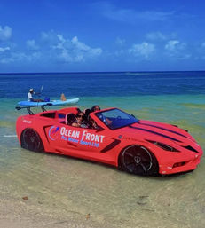 Red convertible sports car parked in shallow turquoise surf on a sunny tropical beach with two passengers smiling and a kayaker paddling nearby under a bright blue sky